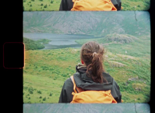 A female hiker with a yellow backpack looks out at a stunning, foggy mountain vista with a lake, captured on vintage film