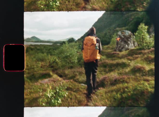 Vintage film scan effect shows a hiker with an orange backpack walking on a trail through a lush, green valley towards a distant lake