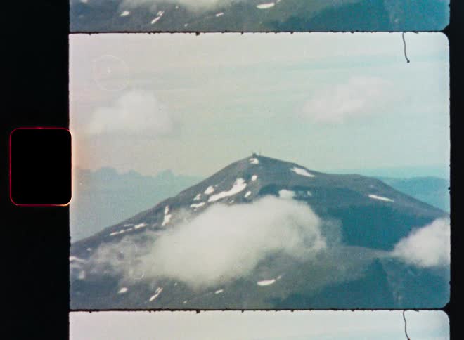 Vintage 8mm film scan of a majestic, snow-dusted mountain seen from an aerial perspective with swirling clouds