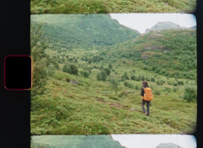 A person wearing a bright orange backpack hikes down a trail in a lush, green mountain valley in this vintage-style 8mm film