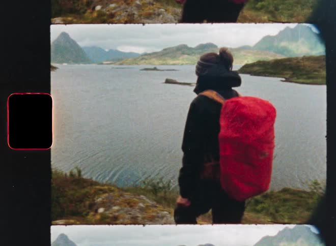 Vintage film footage captures a hiker with a red backpack standing on a windy shore, looking out at a scenic mountain lake