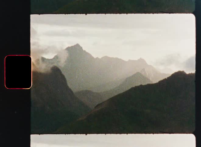 A vintage 8mm film recording slowly zooms in on the misty, cloud-covered peaks of a distant mountain range