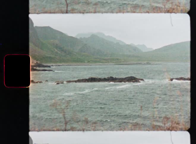 A vintage 8mm film recording slowly zooms in on a rocky outcropping in a choppy sea with green mountains in the background on an overcast day