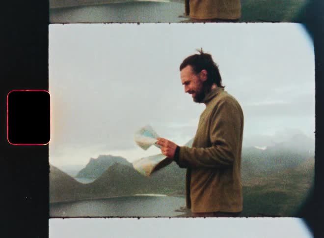 A vintage 8mm film captures a male hiker standing on a windy mountain peak and looking at a map to navigate his route