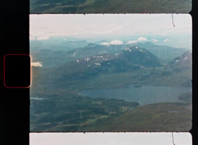 Vintage 8mm film captures an expansive aerial view of a lake nestled among lush green, snow-capped mountains