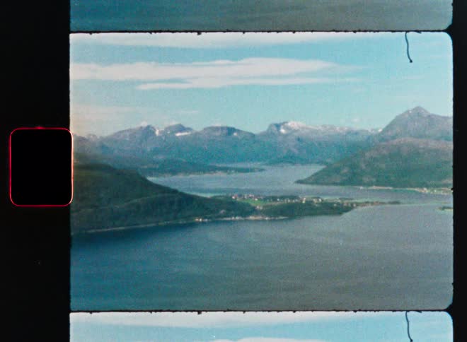 A vintage 8mm film scan reveals a stunning panoramic view of a fjord with snow-capped mountains and a quaint coastal village