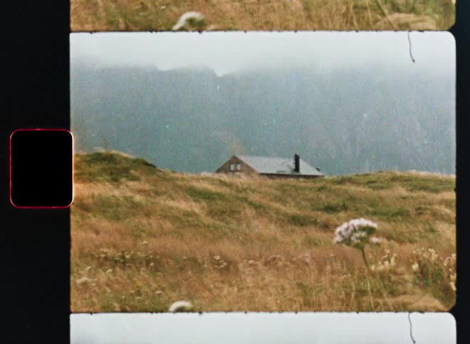 Super 8mm footage captures a lonely cabin on a windy, grassy hill with a hazy mountain backdrop
