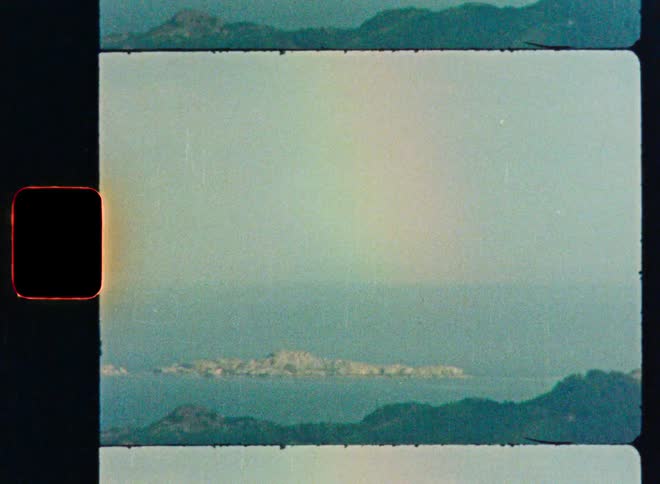 Vintage 8mm film captures a hazy coastal view of a distant island with a faint rainbow in the sky
