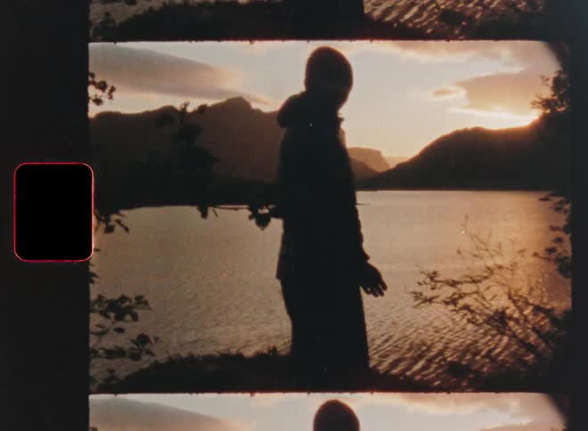 Vintage 8mm film shows a silhouetted person looking out over a mountain lake at sunset