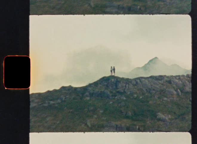 A vintage film shot zooms in on two distant figures standing atop a rugged, green mountain on a cloudy day