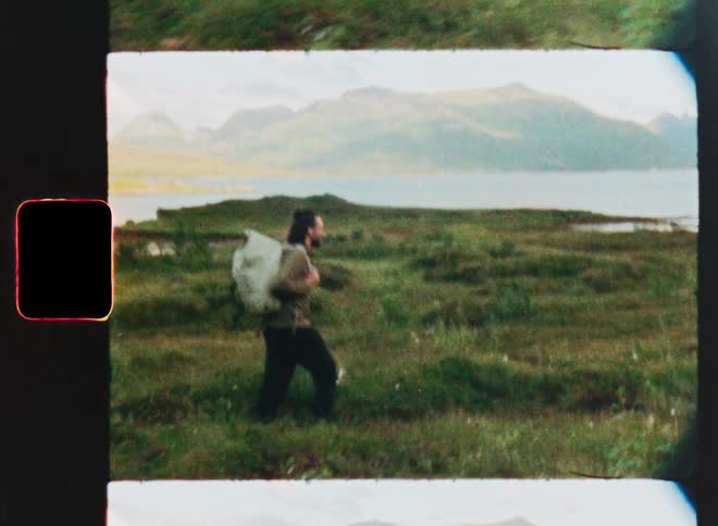 A vintage film scan captures a man with a backpack hiking through a grassy field next to a lake with mountains in the distance
