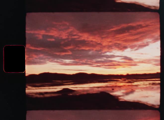 Vintage 8mm footage shows a fiery red and orange sunset reflecting over a tranquil lake or marsh