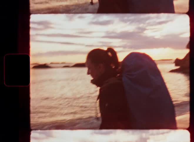 Vintage 8mm film captures a female backpacker standing on a rocky beach at sunset