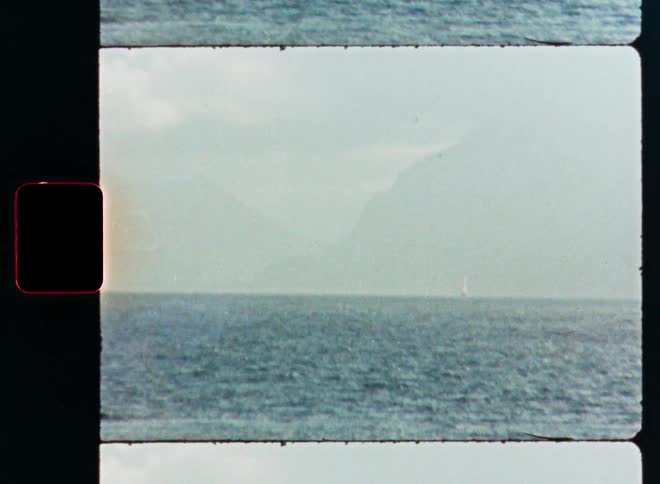 Vintage 8mm film scan shows a tranquil lake with a sailboat in the distance against a backdrop of hazy mountains