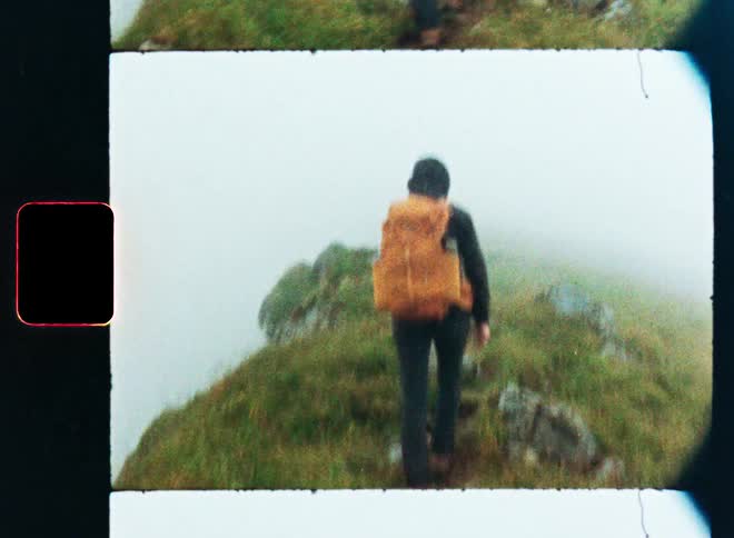 Vintage-style film footage captures a hiker with a backpack walking up a grassy mountain into the fog