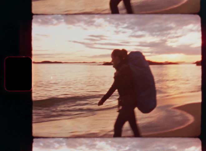 Vintage film footage shows a female backpacker walking along a sandy beach as the sun sets over the water