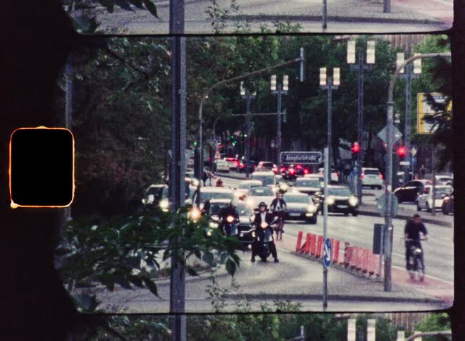 Vintage film footage shows the hustle and bustle of daily traffic on a European city street with cars, motorcycles, and bicycles