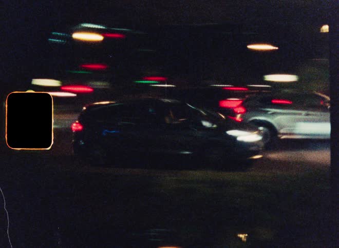 A blurry, grainy, super 8mm film-style shot captures cars speeding down a city street at night, creating vibrant light streaks