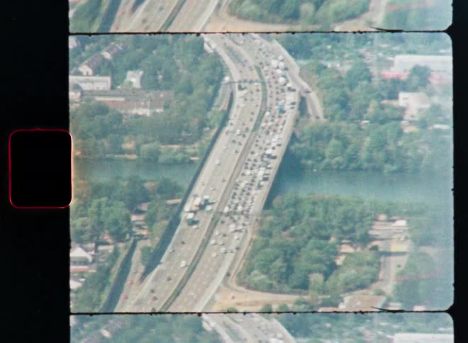 A vintage film aerial shot captures a major highway with a significant traffic jam running alongside a river and through a cityscape