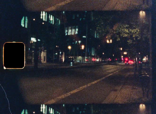 Grainy vintage film captures a time-lapse of city street traffic at night with glowing light trails