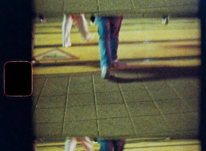 A grainy, vintage film shot captures the reflected, low-angle view of people walking on a city street at night
