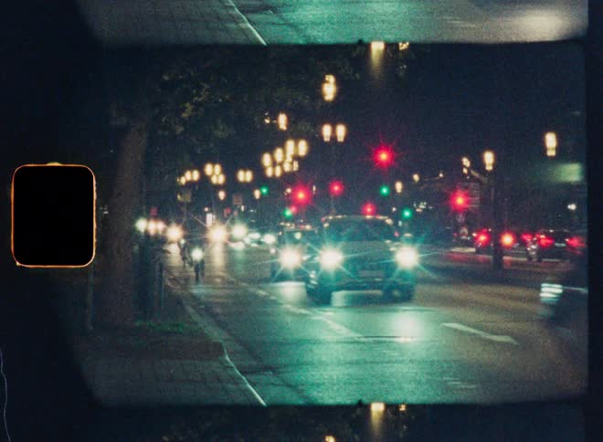 A grainy, vintage film aesthetic captures the vibrant flow of traffic with bright headlights and colorful signals on a busy city street at night