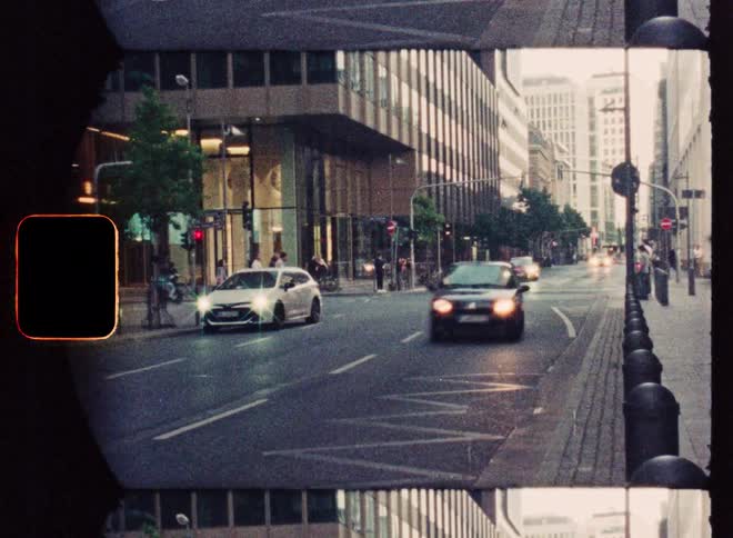 A vintage film strip captures grainy footage of cars driving through a city intersection with pedestrians on the sidewalks