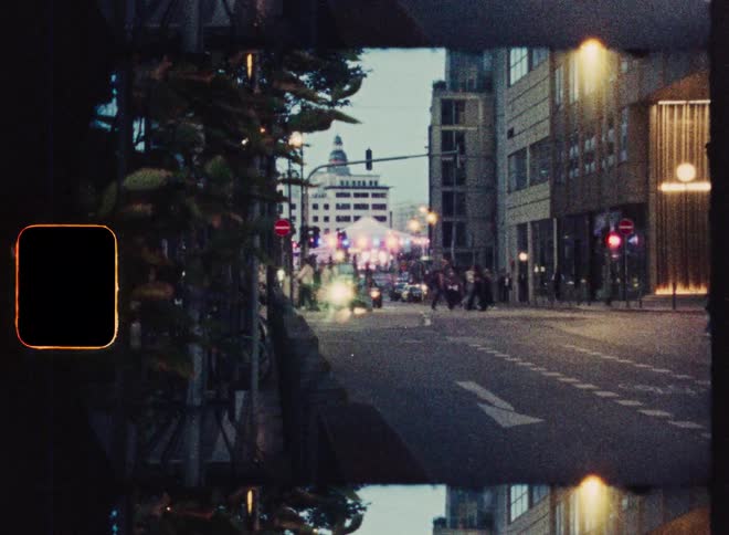 A nostalgic, grainy film view of city life with cars and people moving through an intersection at dusk