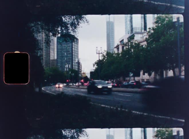 A nostalgic, vintage film-style shot captures the blur of city traffic moving past modern skyscrapers, complete with light leaks and sprocket holes