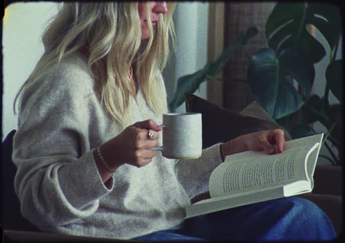 A woman relaxes at home reading a book and holding a mug, filmed with a vintage aesthetic