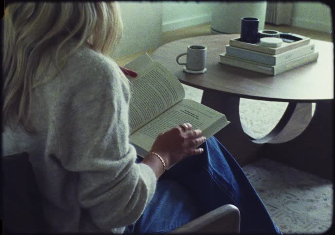 A woman in a cozy sweater sits and reads a book, turning the page, captured with a vintage film aesthetic
