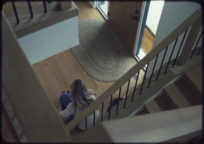 A woman with blonde hair sits at the bottom of a staircase and takes off her boots in a retro-style high-angle shot