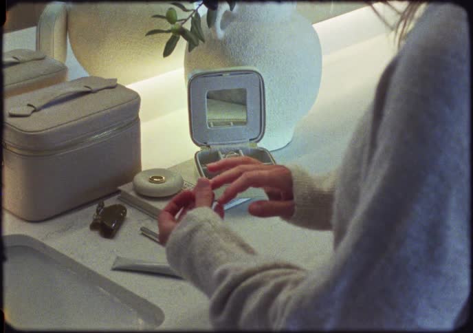 A woman puts on gold rings from her jewelry box while getting ready at her vanity in this vintage-style clip
