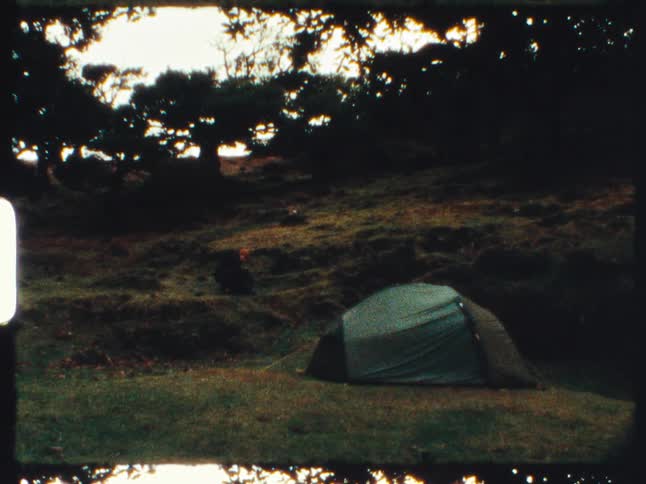 Super 8 film captures a solitary tent pitched in a grassy, wooded clearing as a person rests nearby at dusk