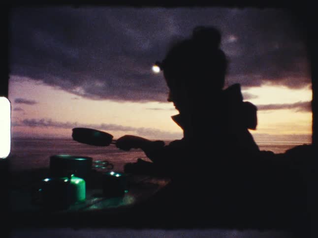 A silhouetted camper with a headlamp cooks a meal on a portable stove against a dramatic sunset sky over the ocean on vintage film