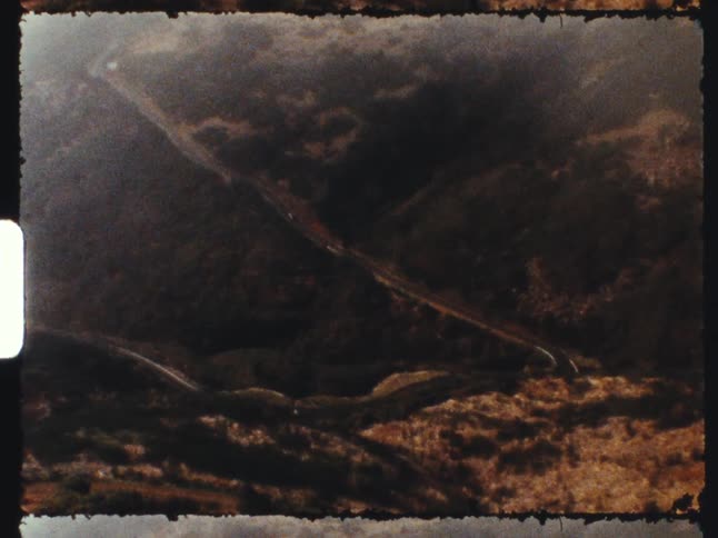Vintage 8mm film captures a hazy mountain landscape with a winding road as shadows move across the valley, ending with a film burn
