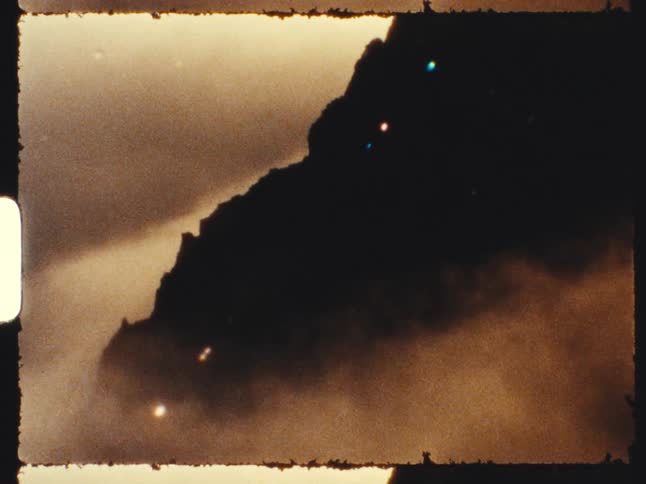 A dark, silhouetted mountain peak emerges from thick, moody fog in a sepia-toned archival film clip
