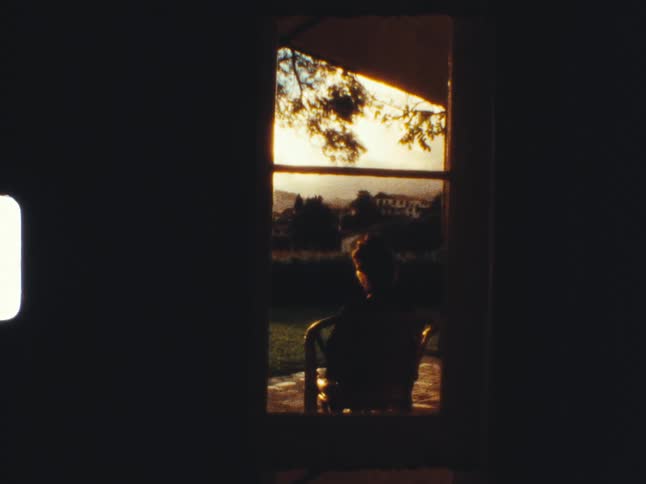 A silhouetted person sits peacefully in a chair on a patio, viewed from a dark room looking out into the warm glow of sunset