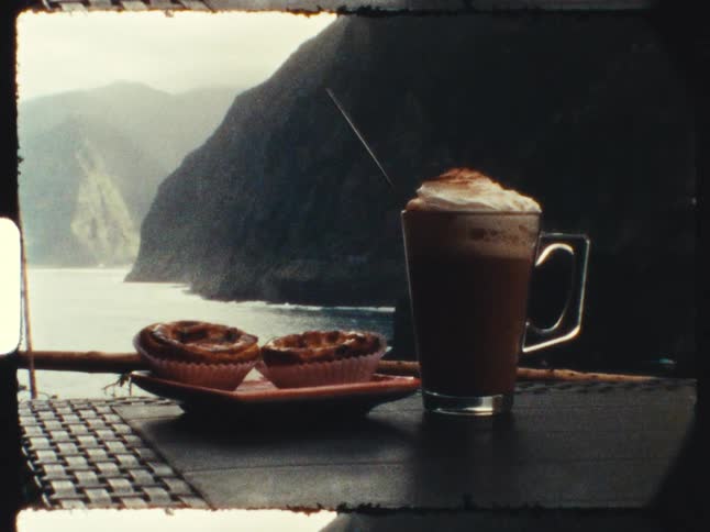 Vintage 8mm film captures a still life of a hot chocolate and Portuguese custard tarts on a table overlooking a dramatic ocean coastline