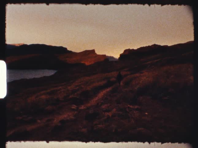 A lone hiker traverses a winding trail through a vast, hilly landscape next to a lake at dusk in this grainy, vintage film shot