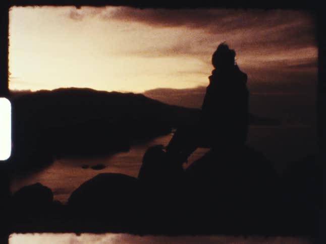 A vintage 8mm film captures the silhouette of a person in a beanie sitting on a rock overlooking a lake and mountains at sunset