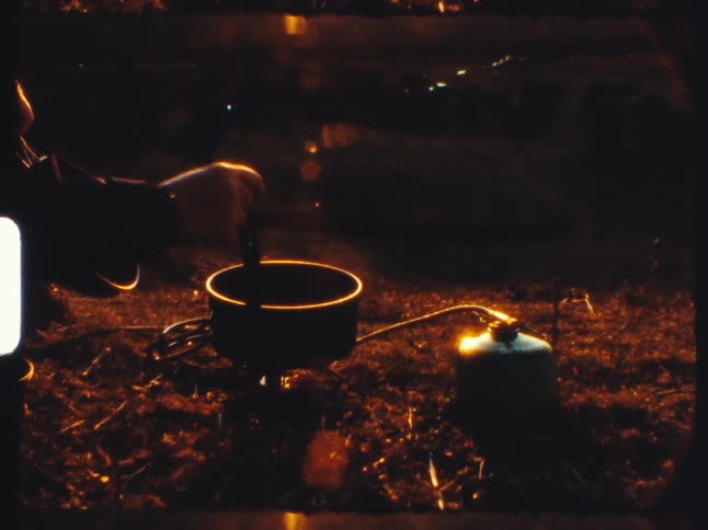 A person cooks a simple meal on a portable camping stove at dusk in this authentic vintage film footage