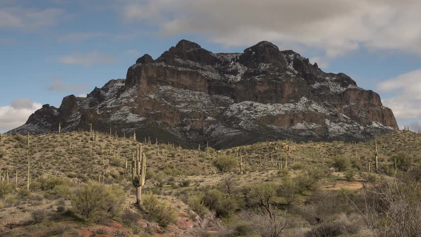 A timelapse of clouds and shadows moving across the iconic Superstition Mountains, which are lightly dusted with snow, rising above the cactus-filled desert