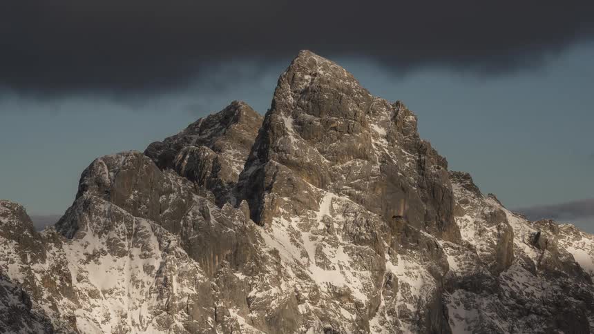 Golden sunset light moves across a majestic, snow-dusted mountain peak as dramatic, dark clouds loom overhead