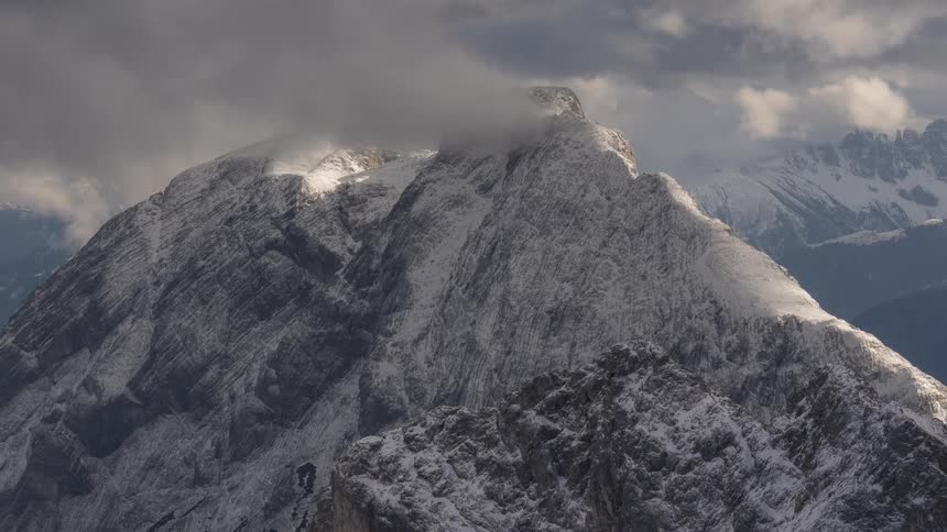 Dramatic sunlight breaks through stormy clouds to illuminate the rugged, snow-covered peaks of a vast mountain range