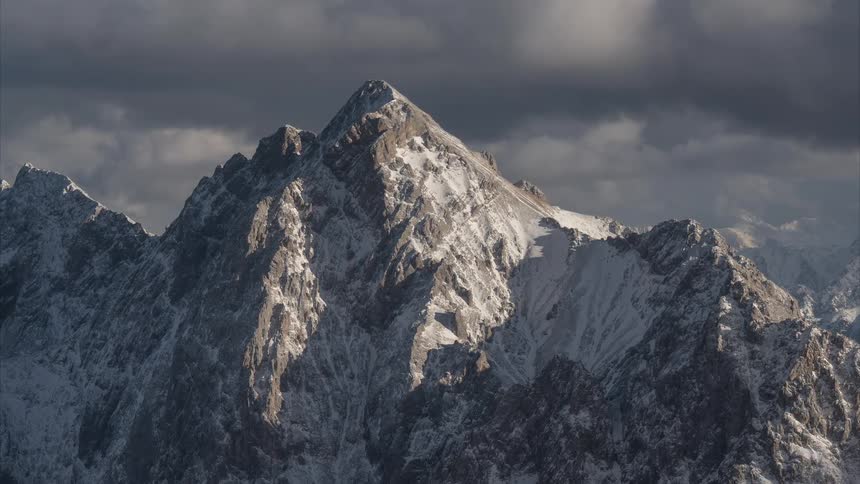 A dramatic time-lapse captures sunlight and shadows dancing across a rugged, snow-covered mountain peak under a stormy sky