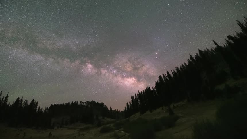 A majestic time-lapse captures the Milky Way galaxy rising over a serene creek flowing through a remote, forested mountain valley