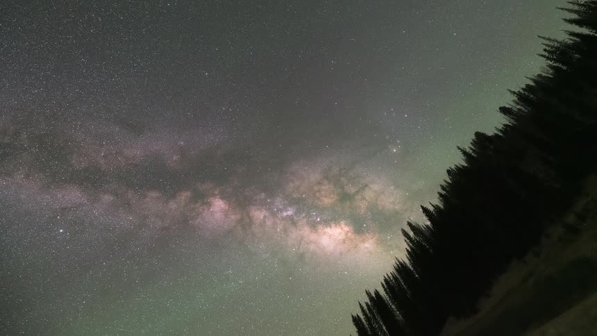 A celestial time-lapse captures the vibrant Milky Way and faint aurora moving across the night sky above a forested mountain valley until dawn
