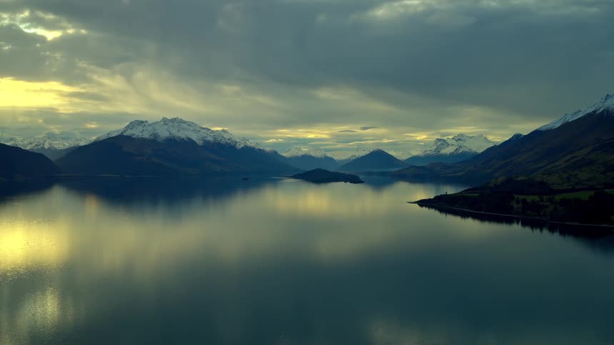 Golden light breaks through dramatic clouds over a still lake, perfectly reflecting the snow-capped mountains