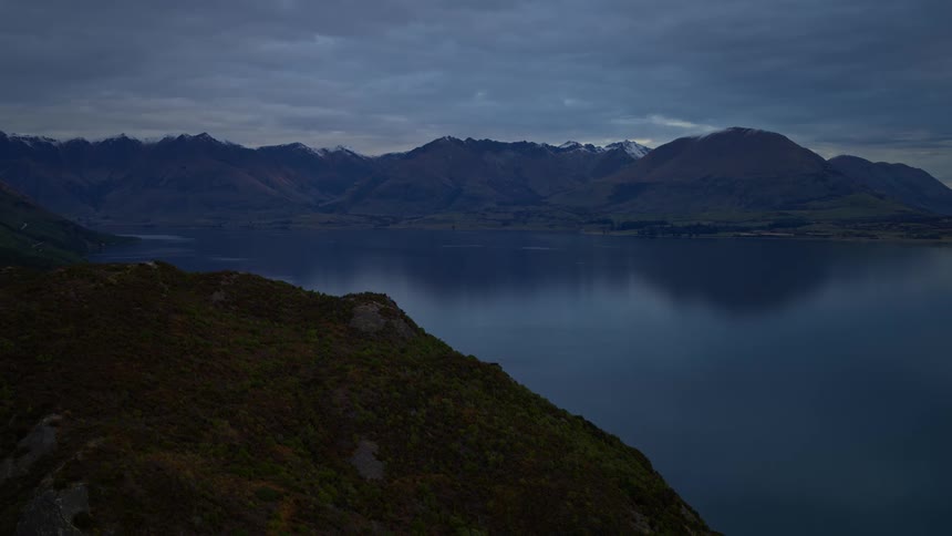 A serene view from a hillside overlooking a tranquil lake reflecting the snow-capped mountains under a dark, cloudy sky at dusk